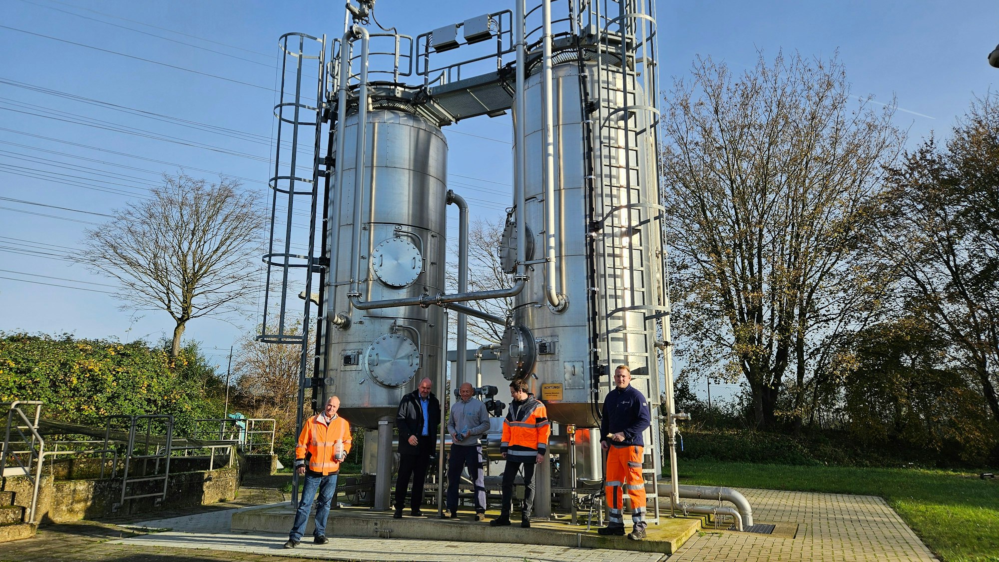 Fünf Männer stehen vor einer Anlage mit zwei silbern glänzenden Silos.