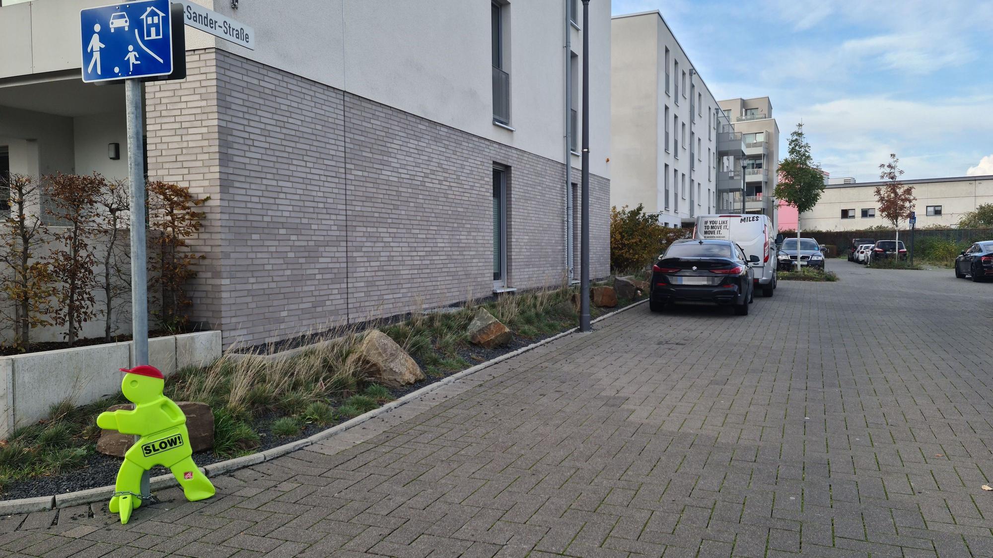Ein Straßenschild mit dem blauen Zusatzschild, das eine Spielstraße auszeichnet und ein gelbes Plastikmännchen stehen an der Einmündung einer Straße, an der auch einige Autos parken.