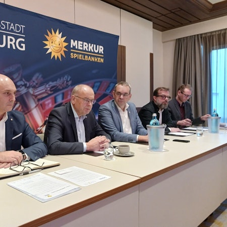 Fünf Mann sitzen an einem langen Tisch vor einem Plakat mit einem Roulette-Tisch