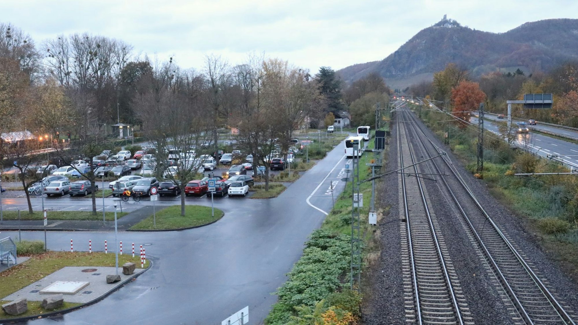 Neben einem Parkplatz verläuft die Bahntrasse, rechts davon befindet sich die Bundesstraße 42. Im Hintergrund ist der Drachenfels zu sehen.