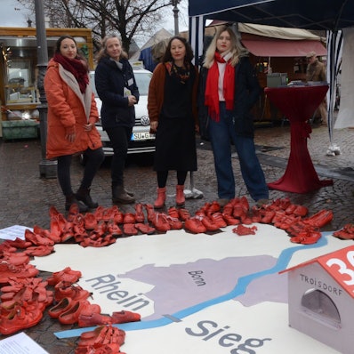 Vier Frauen; sie haben aus roten Schuhen die Umrisse des Rhein-Sieg-Kreises ausgelegt. Im Vordergrund steht ein symbolisches Frauenhaus mit der Aufschrift 30 Betten.