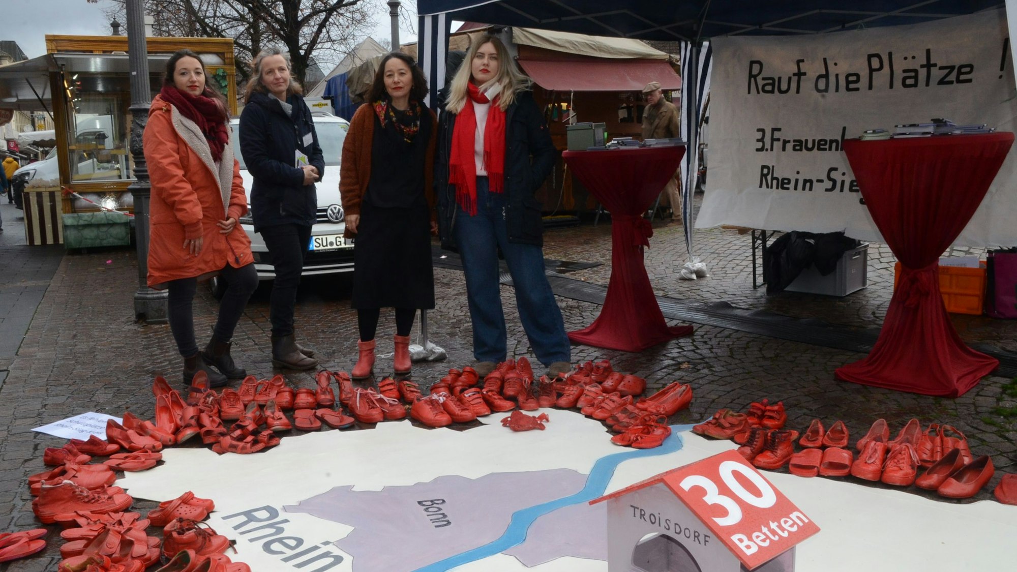 Vier Frauen; sie haben aus roten Schuhen die Umrisse des Rhein-Sieg-Kreises ausgelegt. Im Vordergrund steht ein symbolisches Frauenhaus mit der Aufschrift 30 Betten.