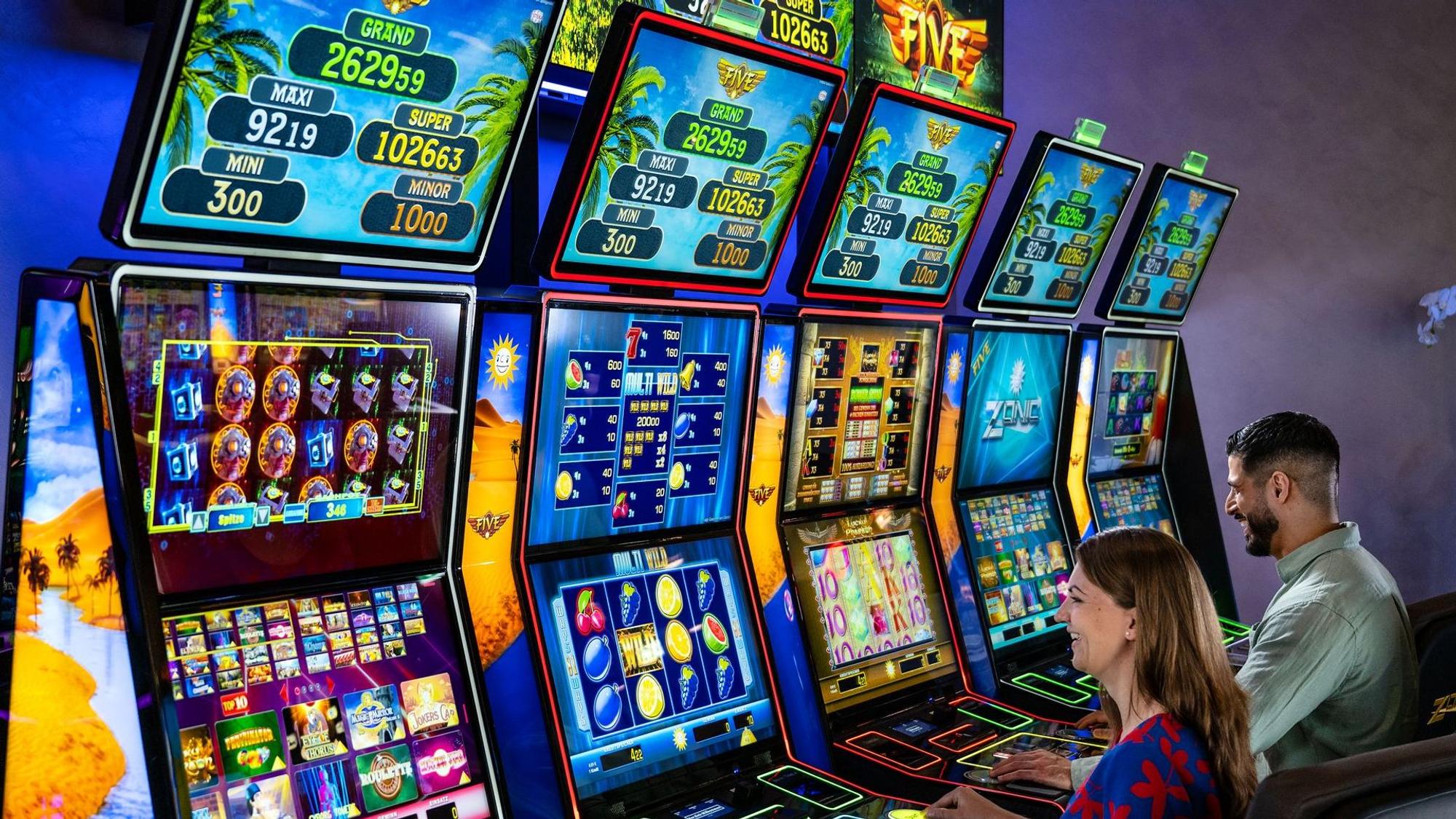 Eine Frau und ein Mann sitzen vor bunten Geldspielautomaten.