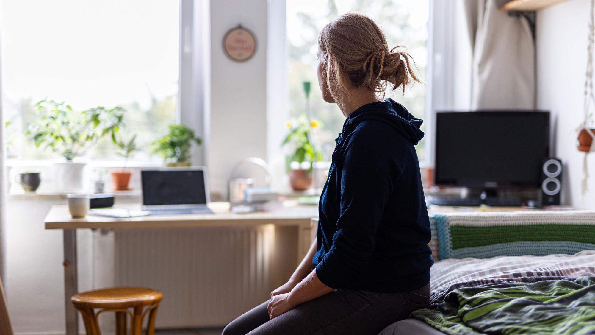 ARCHIV - 13.08.2023, Niedersachsen, Hannover: ILLUSTRATION - Eine Studentin sitzt auf dem Bett in ihrem WG-Zimmer und schaut aus dem Fenster (gestellte Szene).