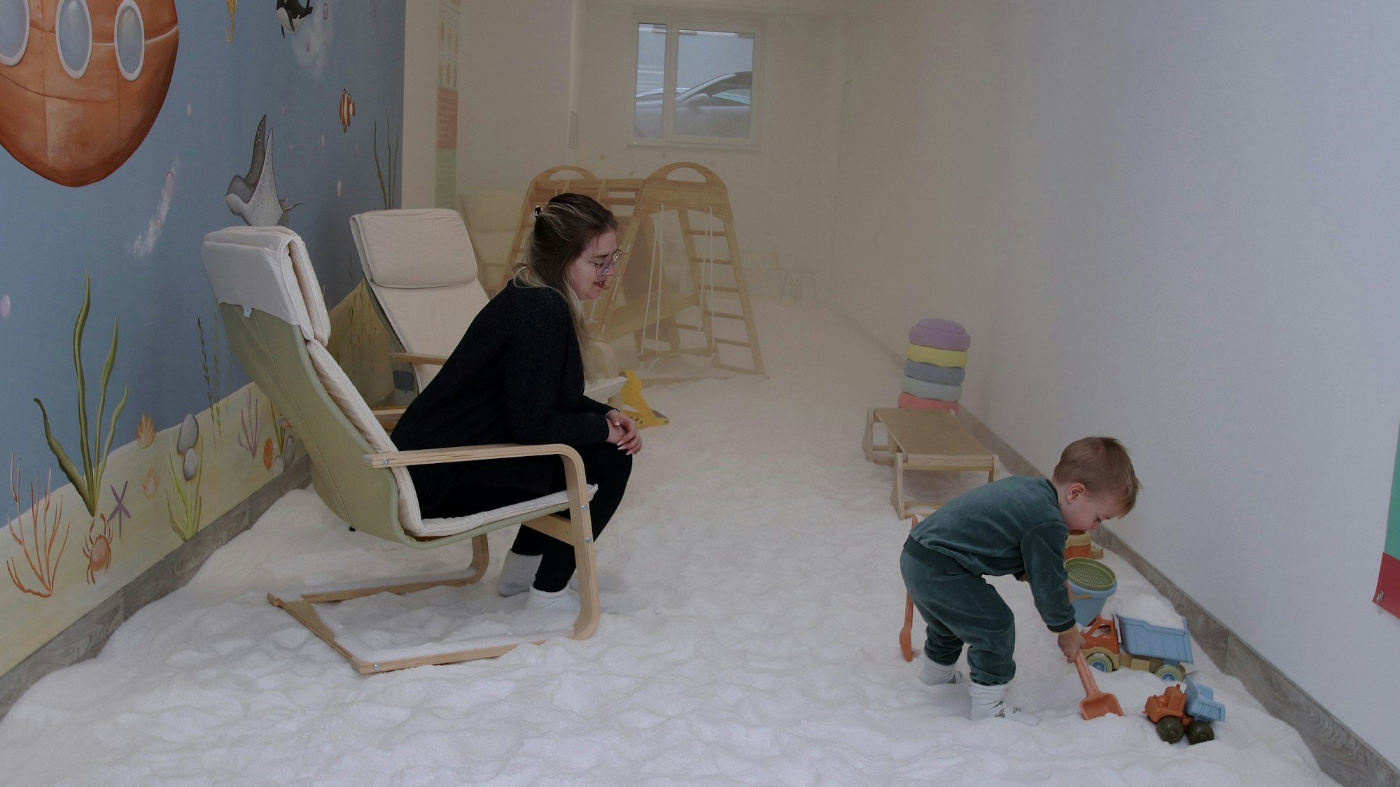 Eine Frau sitzt in einem Sessel, ihr Kind spielt in dem Salzraum.