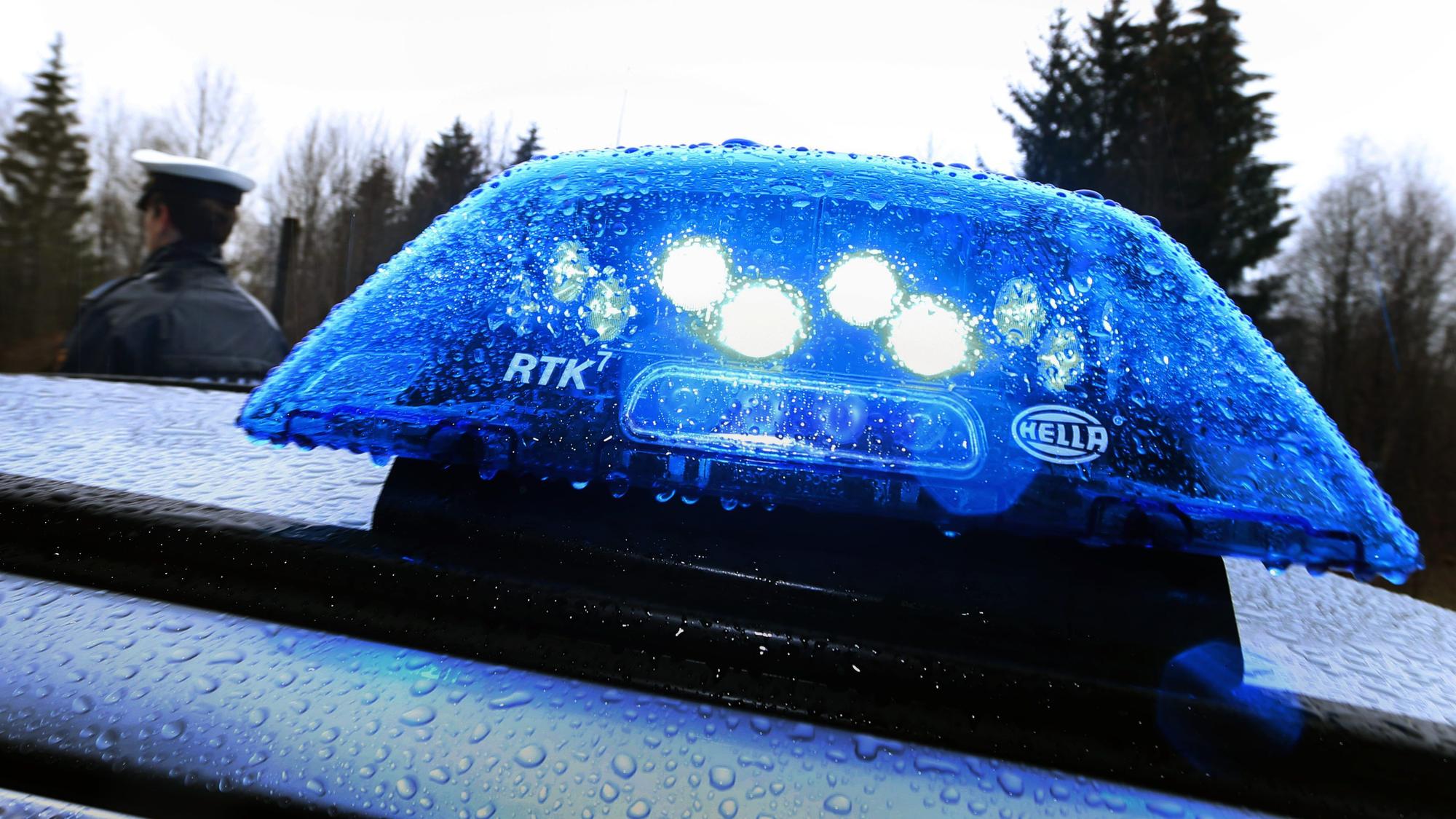 Ein Polizist steht im Regen vor einem Streifenwagen dessen Blaulicht aktiviert ist.