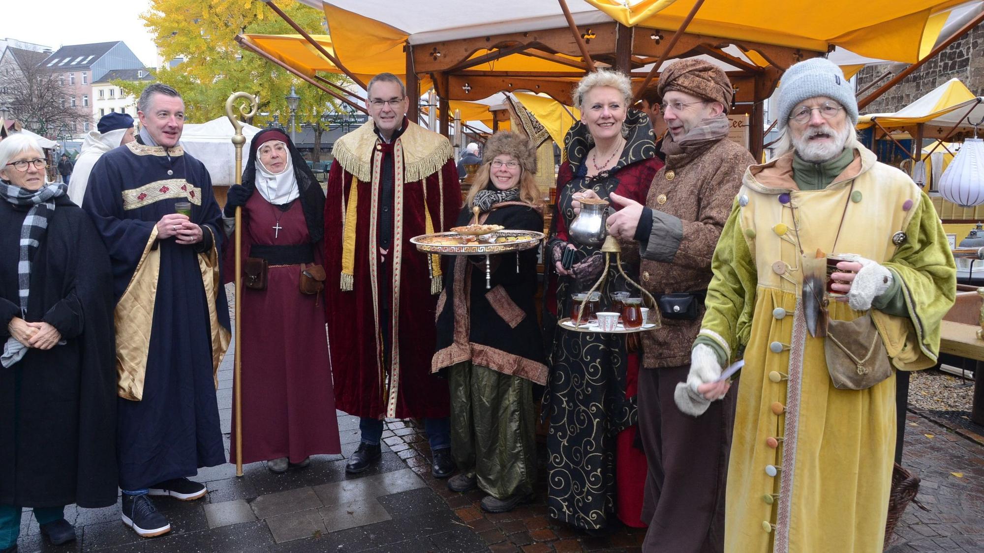 Männer und Frauen in mittelalterlicher Kleidung; eine Frau serviert Fingerfood auf einem Tablett, ein orientalisch gekleideter Mann hat ein Tablett mit Teegläsern in der Hand.