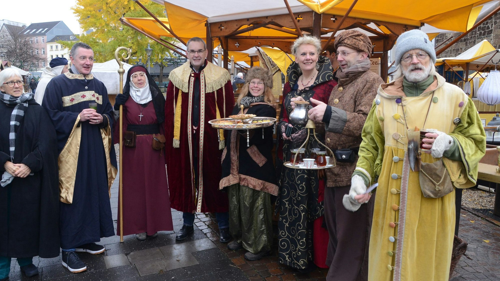 Männer und Frauen in mittelalterlicher Kleidung; eine Frau serviert Fingerfood auf einem Tablett, ein orientalisch gekleideter Mann hat ein Tablett mit Teegläsern in der Hand.