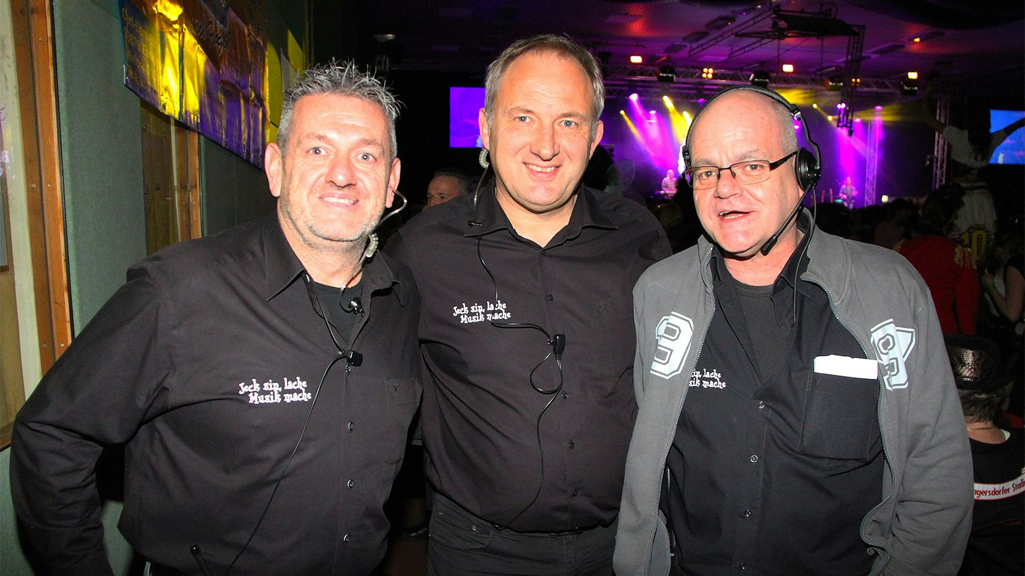 Drei Männer in schwarzen Hemden stehen nebeneinander und lächeln in die Kamera.