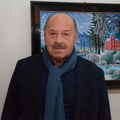 Ein Mann mit Schal und schwarzem Sakko steht vor einem naiven Gemälde.In einer Winterlandschaft leuchtet das rote Herrenhaus der Troisdorfer Burg Wissem..