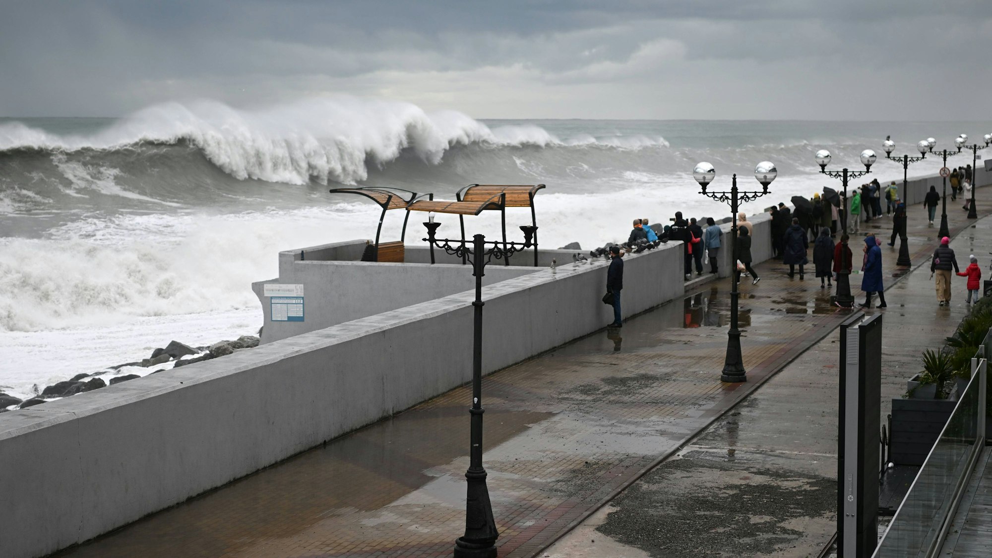 Menschen beobachten eine große Welle, die im russischen Sotschi auf Land trifft. IMAGO/SNA