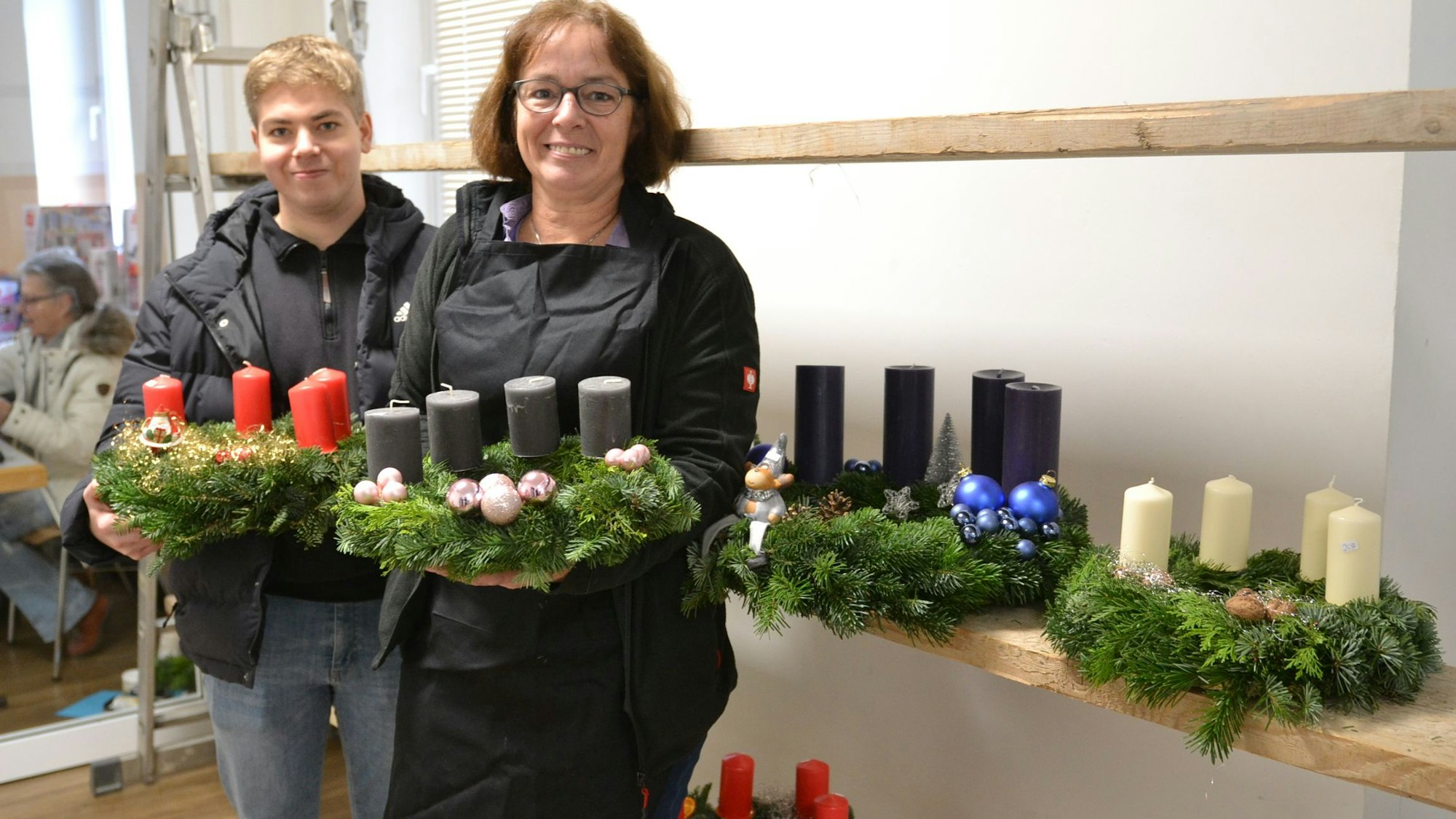 Mutter Michaela Lingscheid und Sophn Stefan halten jeweils einen Adventskranz in den Händen, im Hintergrund weitere Kränze.