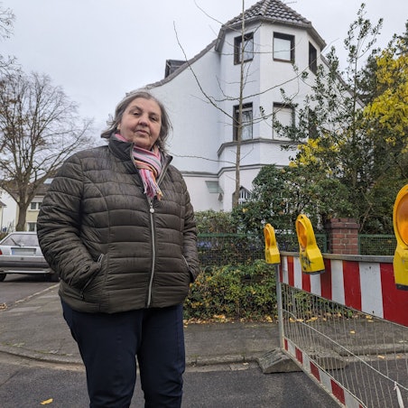 Eine Frau steht neben einer Baustellenabsperrung, im Hintergrund ist ihr Wohnhaus zu sehen.