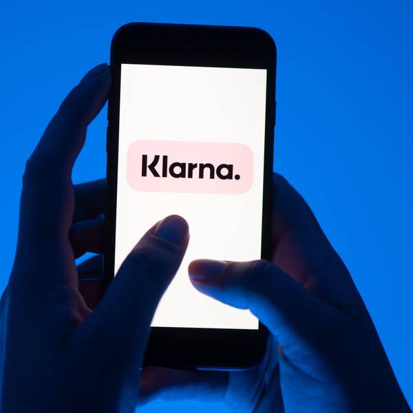 Zwei Hände halten ein Smartphone vor blauem Hintergrund, darauf ist groß das Klarna-Logo zu sehen.