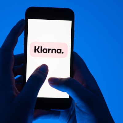 Zwei Hände halten ein Smartphone vor blauem Hintergrund, darauf ist groß das Klarna-Logo zu sehen.