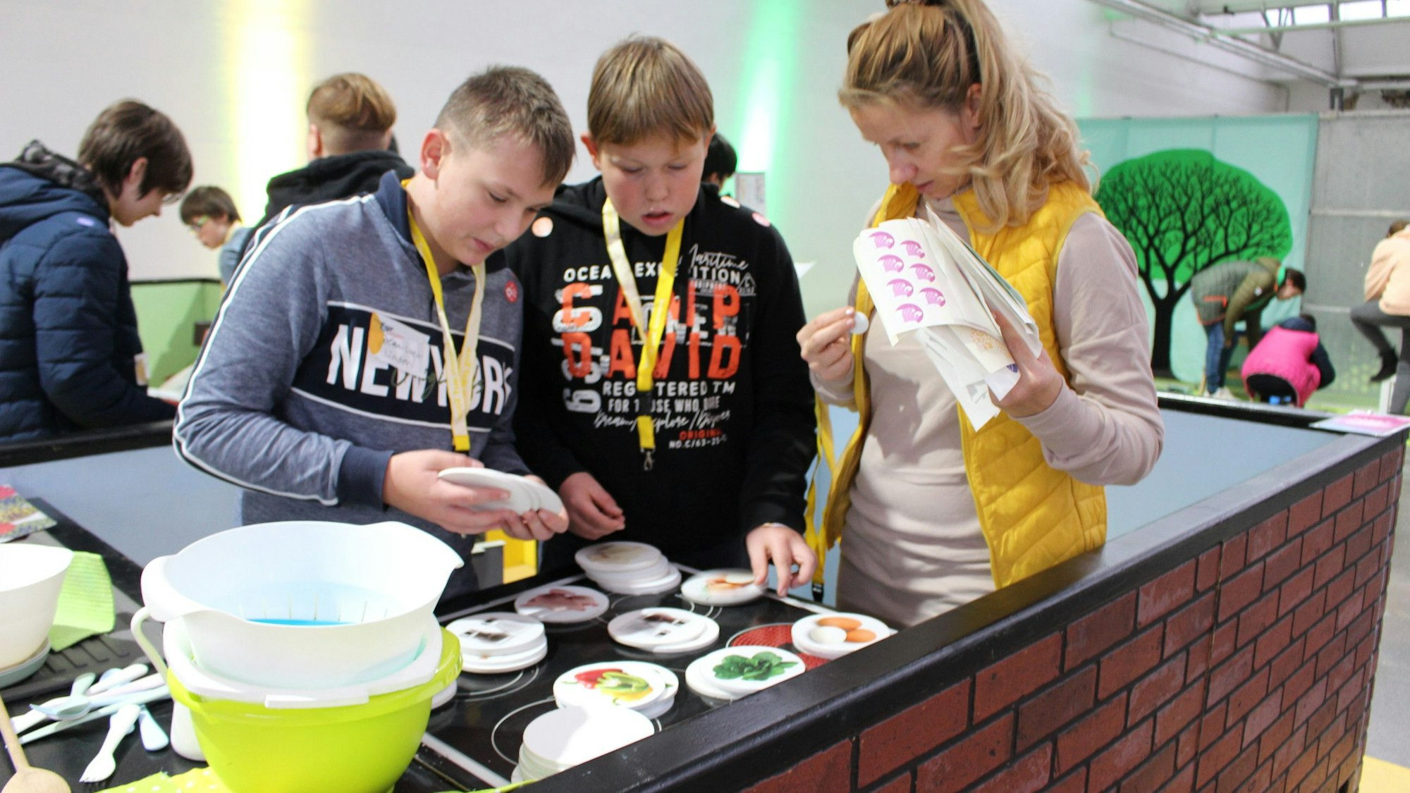 Zwei Schüler räumen in einer Modellküche auf und sortieren Bilder von Lebensmitteln. Eine Begleiterin steht daneben und bereitet die Vergabe eines Stärke-Aufklebers für Kreativität vor.