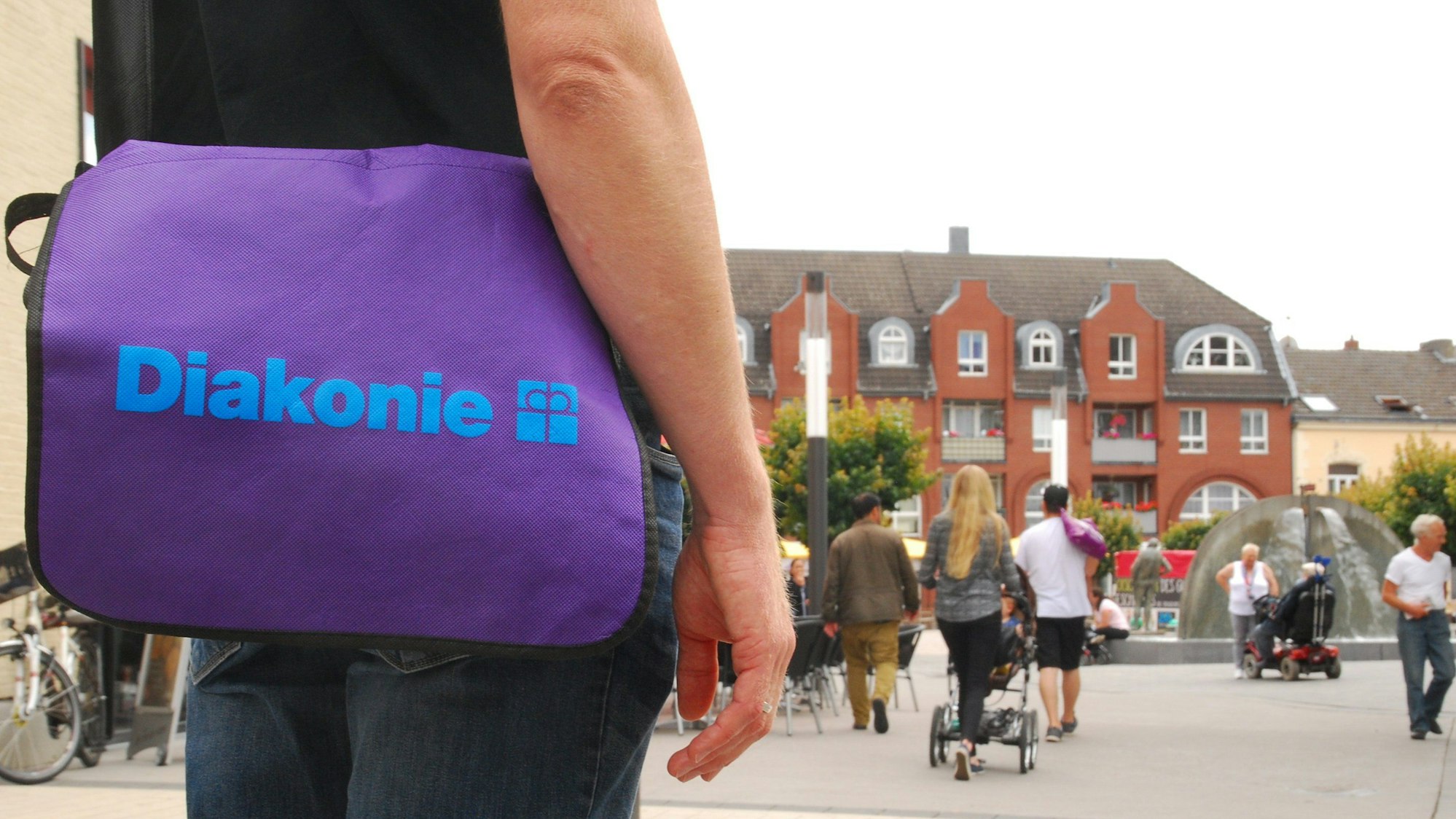 Ein Mann trägt eine violette Umhängetasche mit der Aufschrift Diakonie. im Hintergrund sind Menschen in der Troisdorfer Fußgängerzone zu sehen.
