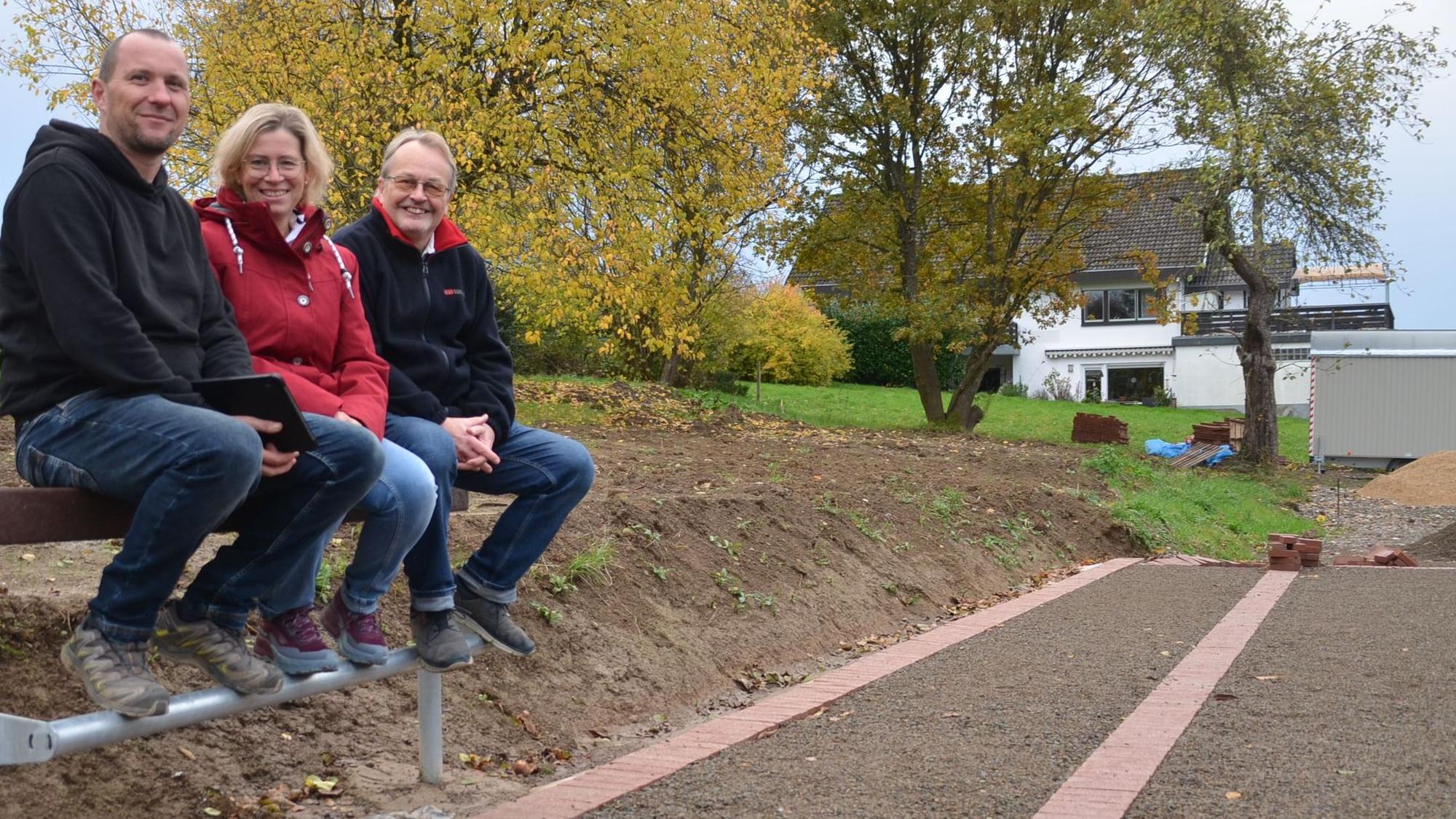 Sie fühlen sich wohl auf der Jugendbank und vor allem auf dem neuen Dorfplatz mitten in Waldbröl-Rossenbach (von links): Landschaftsgärtner und Chefplaner Tobias Pinger sowie Katja Grunewald und Georg Brenner vom Vorstand des Dorfvereins. Die Drei sitzen auf einer der neuen Jugendbänke an dem Platz.