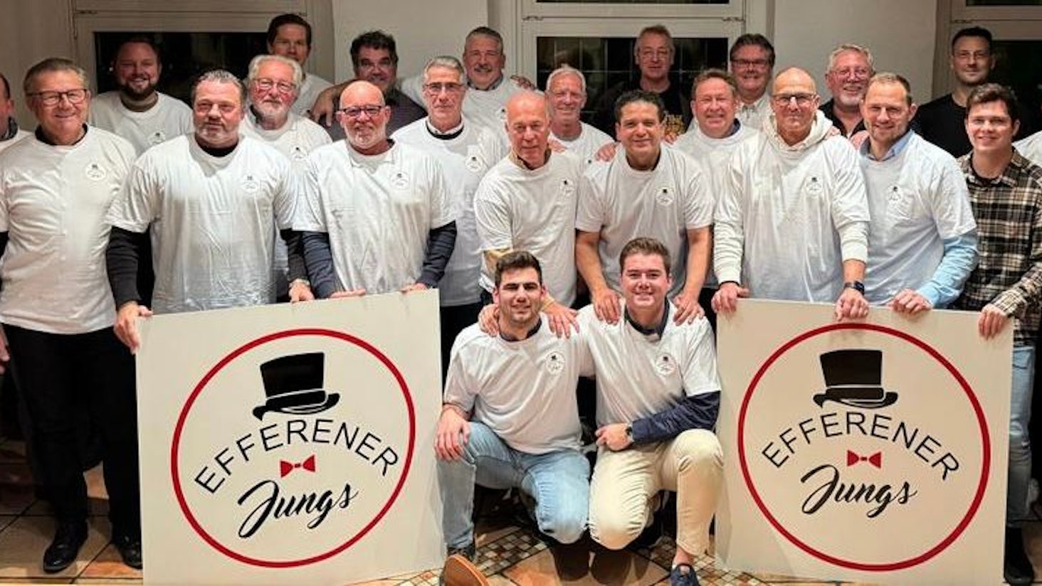 Das Foto zeigt eine Gruppe von Männern in weißen T-Shirts mit zwei großen Schildern mit dem Vereinslogo.
