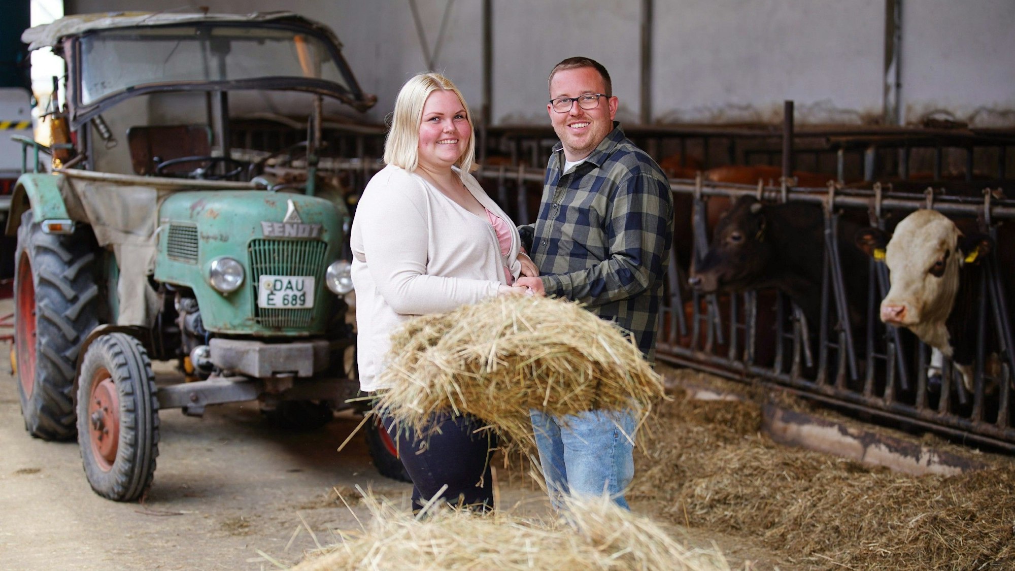Laura und Stephan halten gemeinsam eine Gabel mit Heu, im Hintergrund stehen ein alter Traktor und einige Rinder.