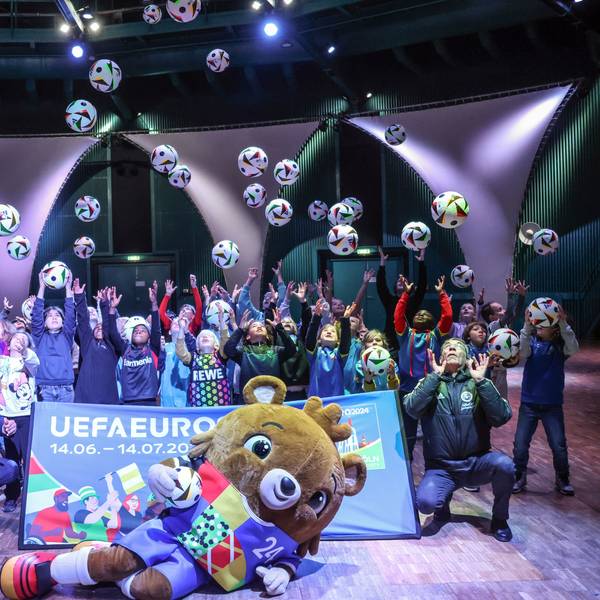 Kinder werfen Bälle in die Luft, das EM-Maskottchen liegt vor ihnen und einem Banner auf dem Boden, auch zwei Erwachsene sind dabei.