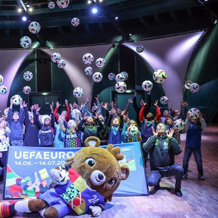 Kinder werfen Bälle in die Luft, das EM-Maskottchen liegt vor ihnen und einem Banner auf dem Boden, auch zwei Erwachsene sind dabei.