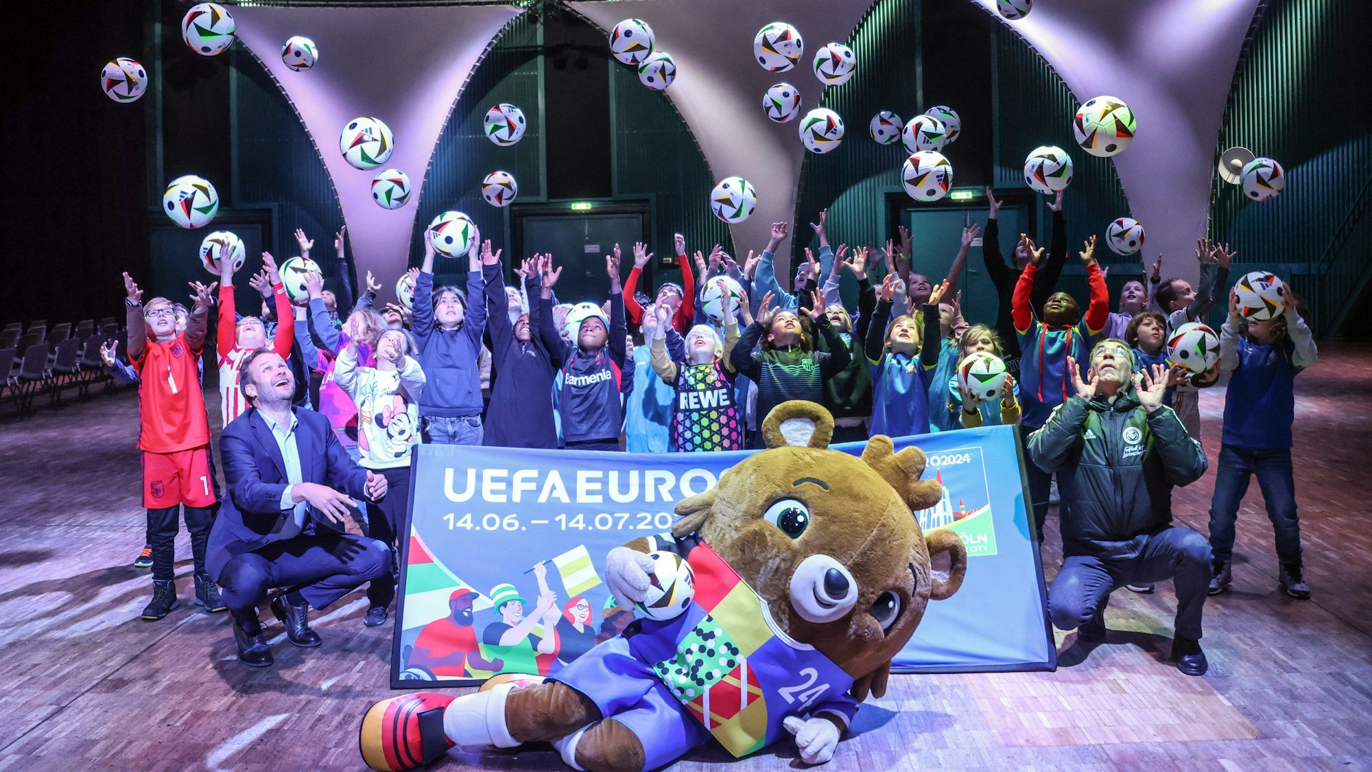 Kinder werfen Bälle in die Luft, das EM-Maskottchen liegt vor ihnen und einem Banner auf dem Boden, auch zwei Erwachsene sind dabei.