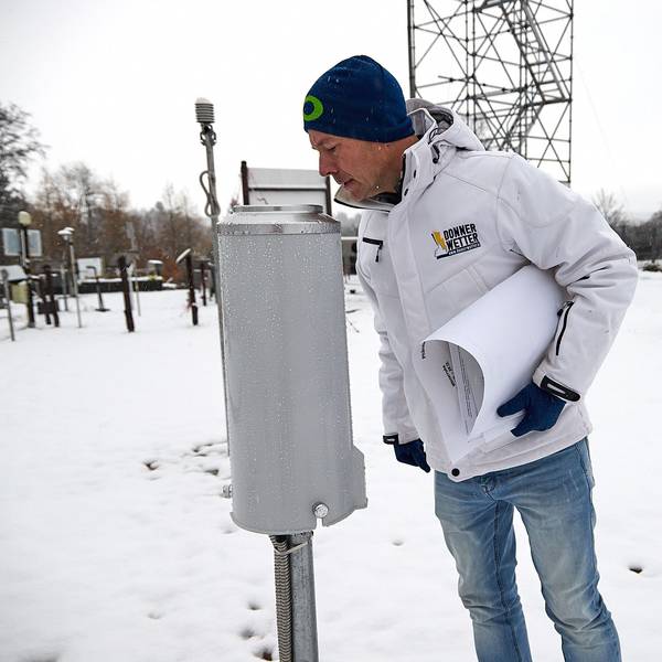 Im Vordergrund schaut Dr. Karsten Brandt in das zylindrische Niederschlagsmessgerät, im Hintergrund das Gerüst der verschneiten Wetterstation.