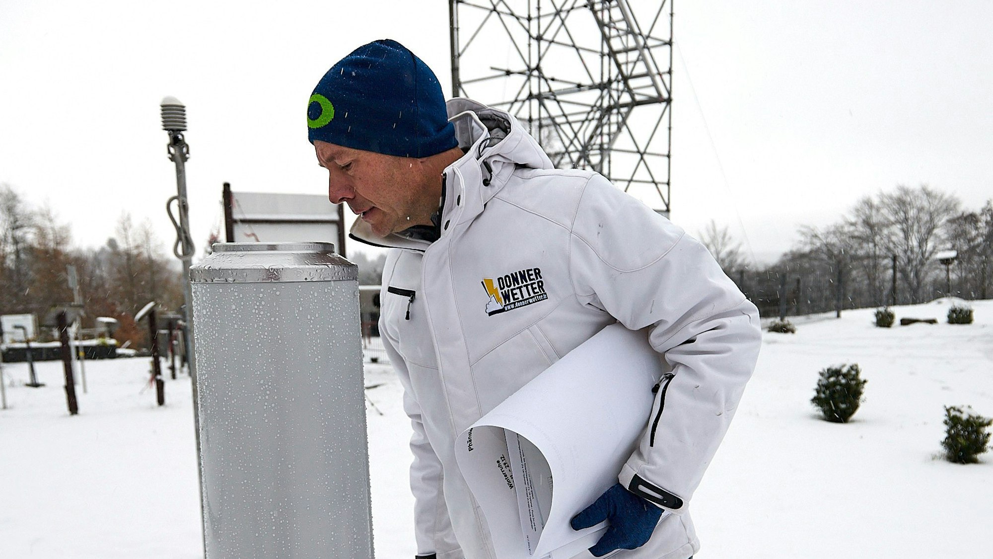 Im Vordergrund schaut Dr. Karsten Brandt in das zylindrische Niederschlagsmessgerät, im Hintergrund das Gerüst der verschneiten Wetterstation.