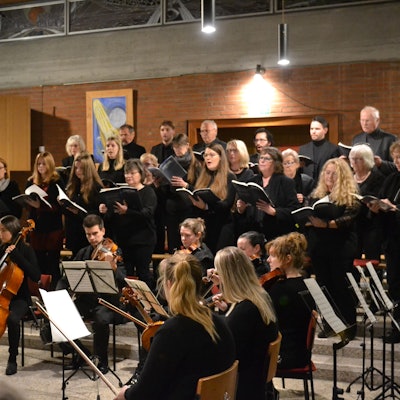 Ein Chor singt in schwarzer Kleidung, davor sitzt ein keines Orchester.