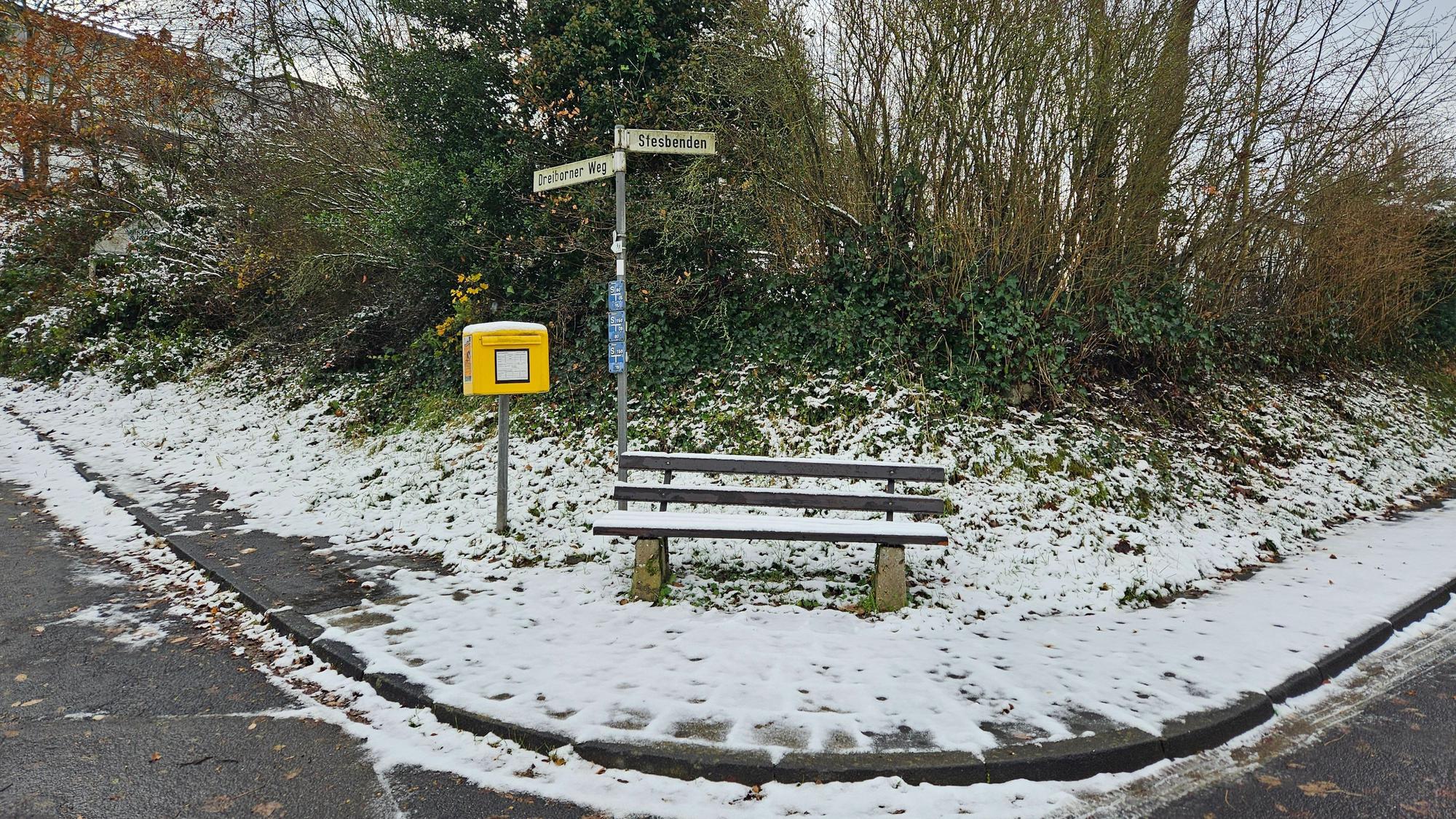 Zu sehen ist eine verschneite Bank mit Briefkasten der Post an einer Straßenkreuzung.