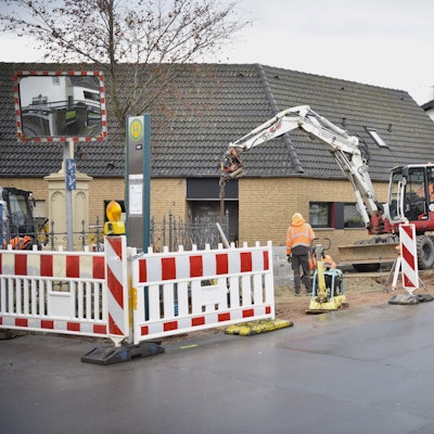 Arbeite in Orangen Jacken stehen an einem abgesperrten Stück Straße, ein Bagger und ein Lastwagen sind ebenfalls zu sehen.