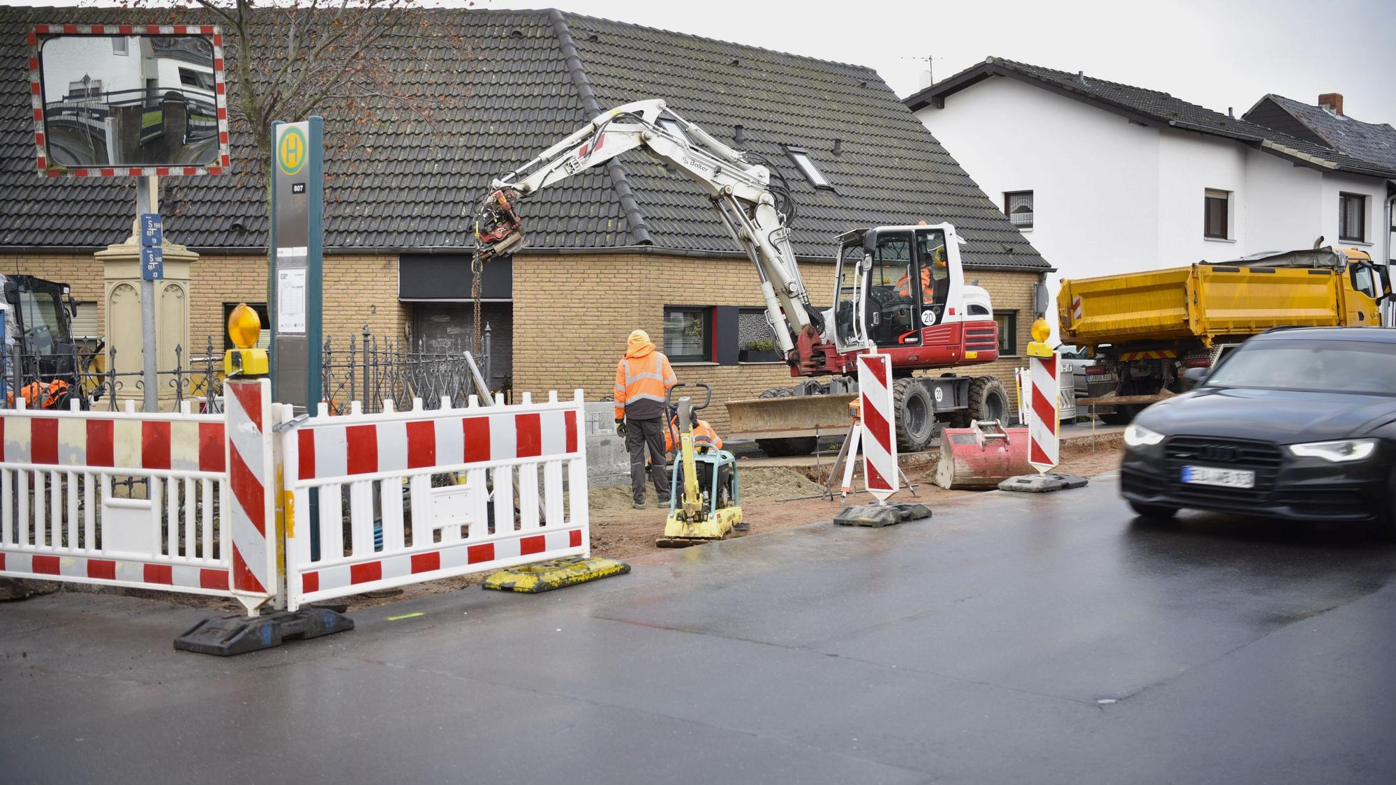 Arbeite in Orangen Jacken stehen an einem abgesperrten Stück Straße, ein Bagger und ein Lastwagen sind ebenfalls zu sehen.