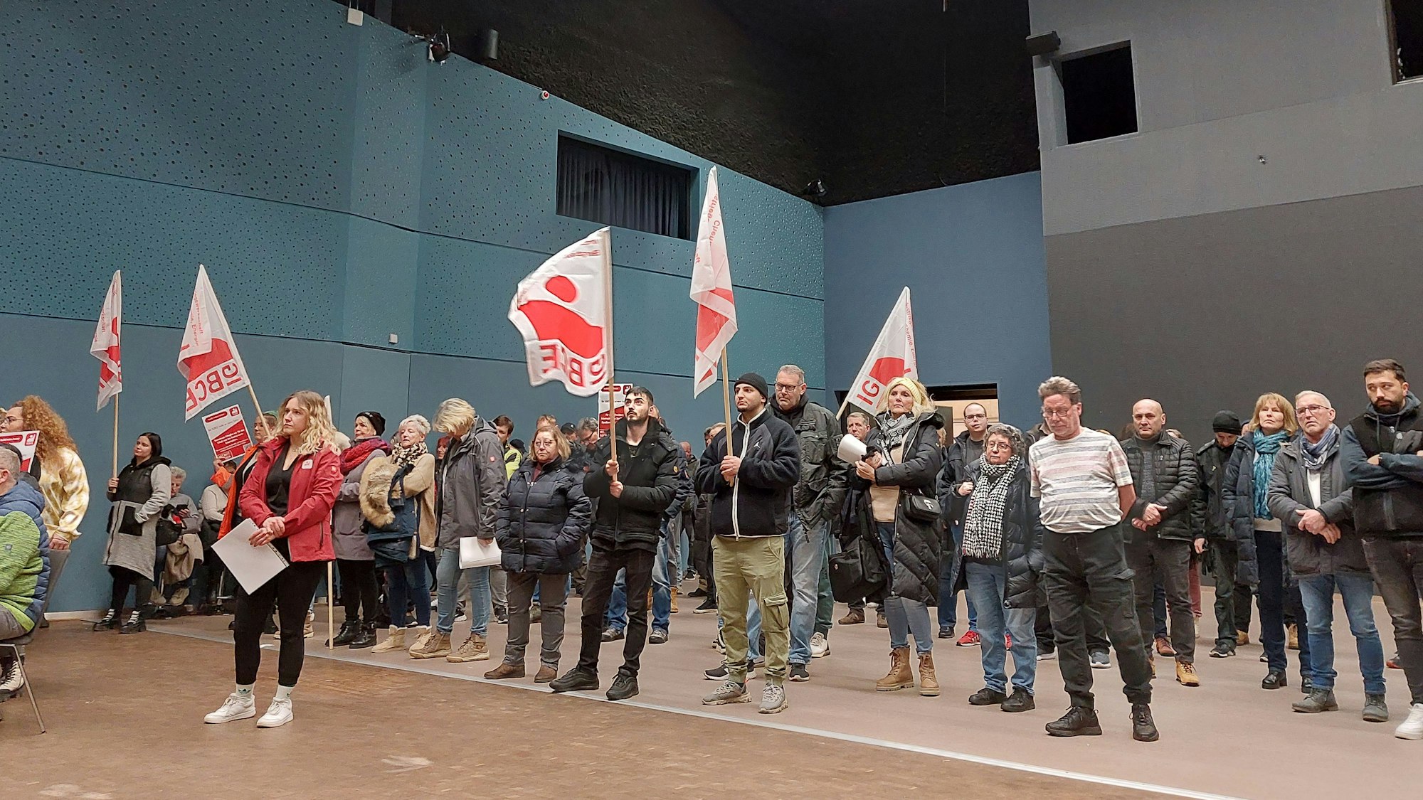 Männer und Frauen stehen in einer Veranstaltungshalle. Einige haben weiß-rote Fahnen ihrer Gewerkschaft dabei. Die Stimmung ist ernst.