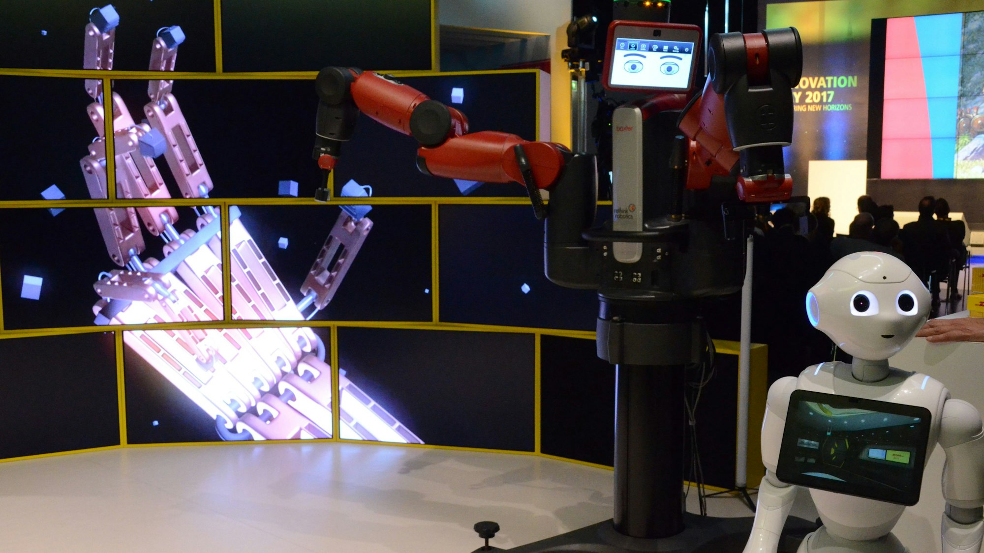 Zwei menschenähnliche Roboter vor einer Videowand aus mehreren Bildschirmen.