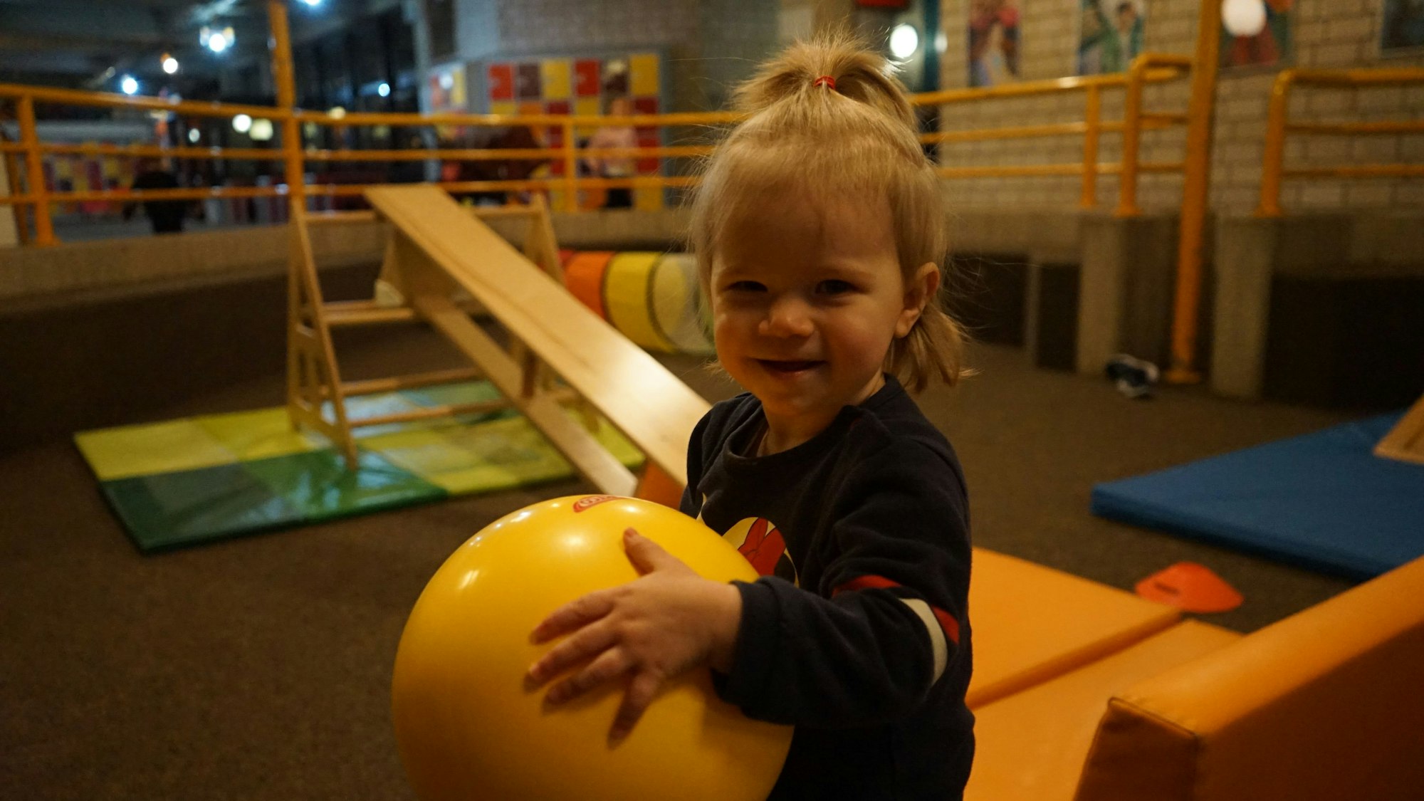 Das Bild zeigt ein Mädchen mit einem gelben Gummiball, im Hintergrund ist ein Bewegungsparcours aus Matten und Sporthallenbänken aufgebaut.