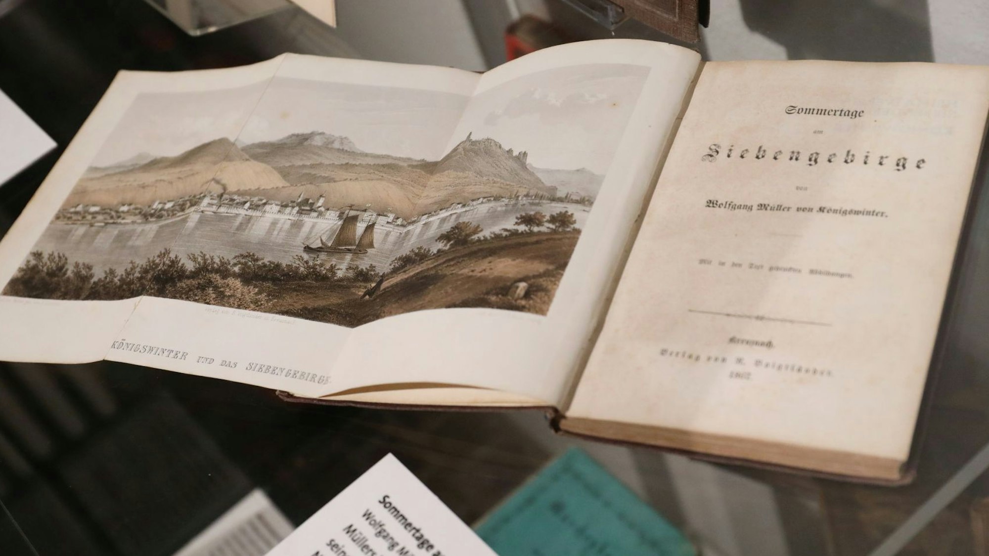 Ein aufgeschlagenes Buch mit dem Titel „Siebengebirge“ zeigt eine Ansicht der Landschaft mit Drachenfels und Rhein.