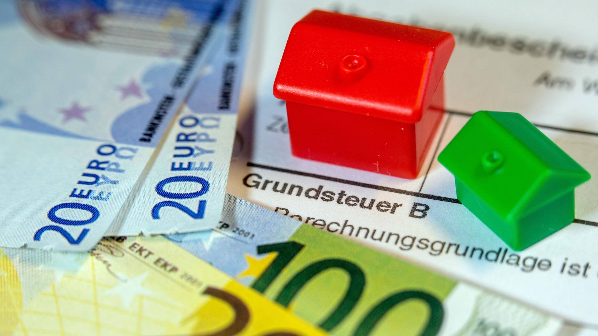 Zu sehen sind Geldscheine, kleine Häuser aus einem Spiel und darunter der Schriftzug Grundsteuer B.