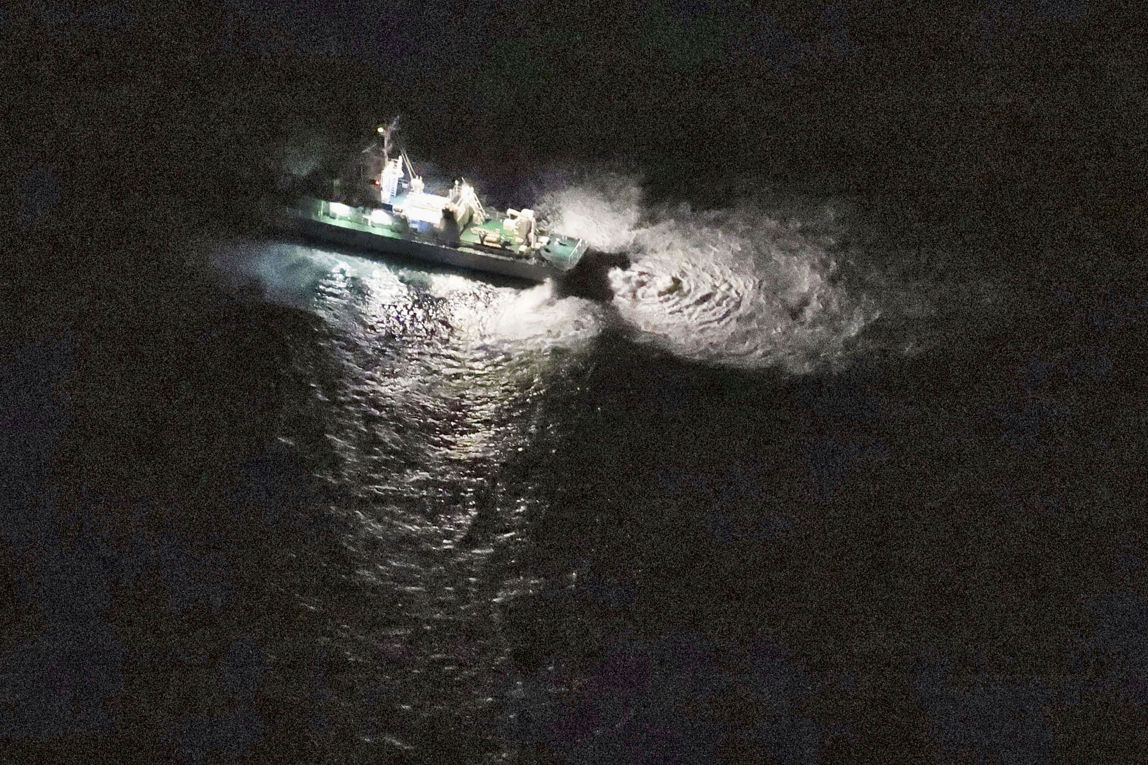 Eine Luftaufnahme zeigt ein Schiff der Küstenwache, dass auf dunkles Wasser leuchtet, in dem dadurch Wellen erkennbar werden.