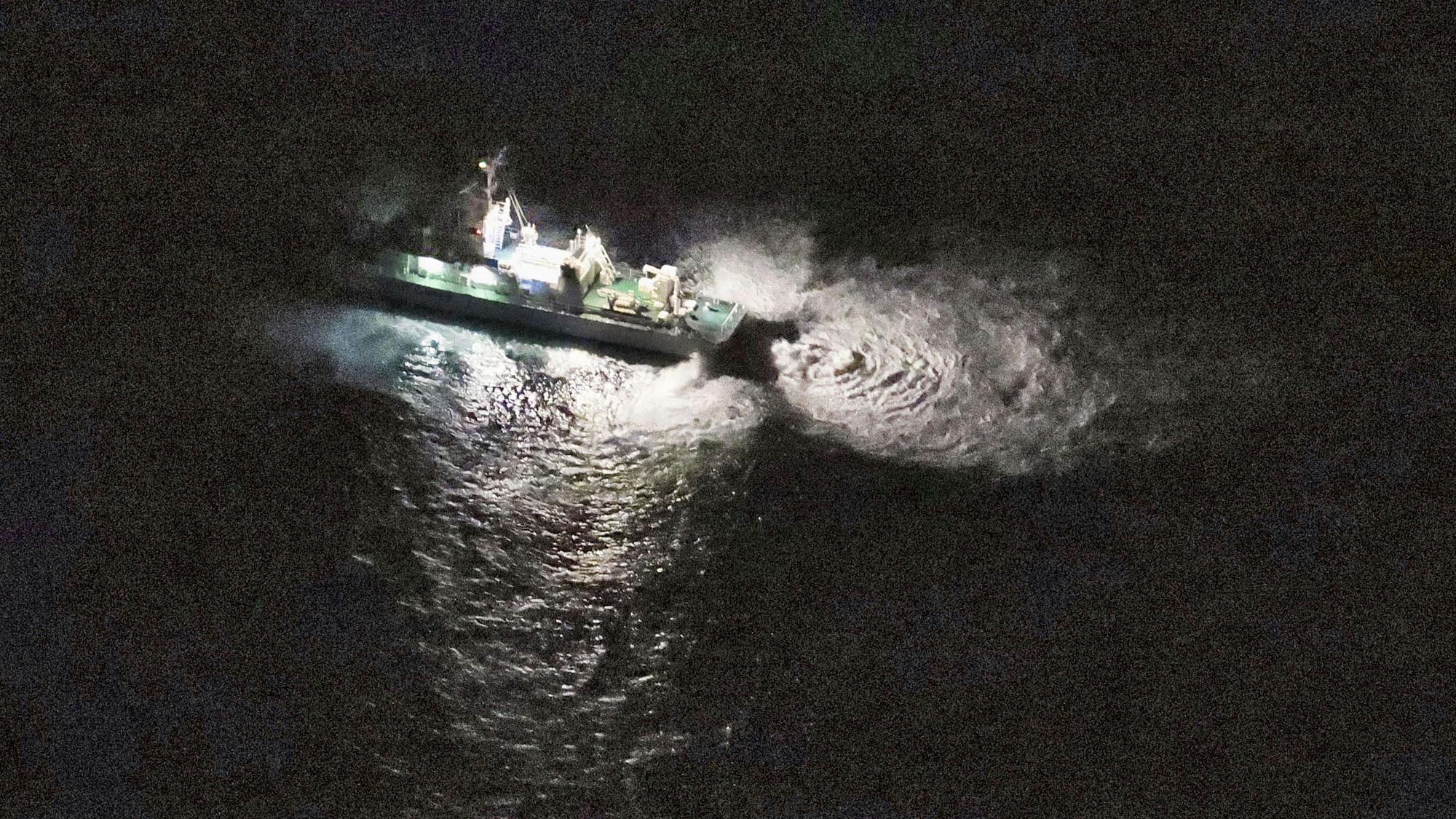 Eine Luftaufnahme zeigt ein Schiff der Küstenwache, dass auf dunkles Wasser leuchtet, in dem dadurch Wellen erkennbar werden.