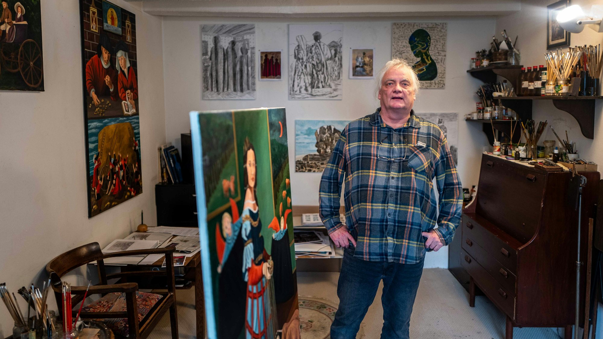Michael Münch steht in seinem Atelier, an den Wänden hängen Bilder, in einer Ecke steht eine Kommode, auf der viele Pinsel und Malutensilien lagern.