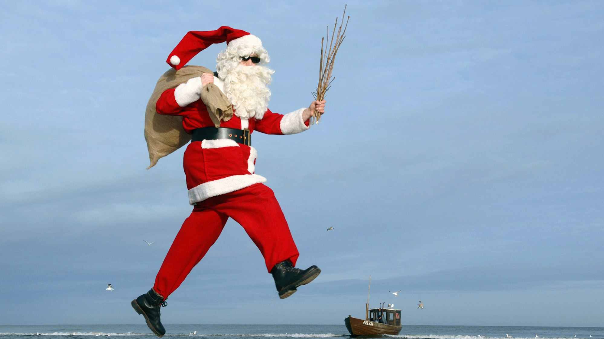 Ein Mann im Nikolauskostüm mit Sonnenbrille hüpft an einem Strand.