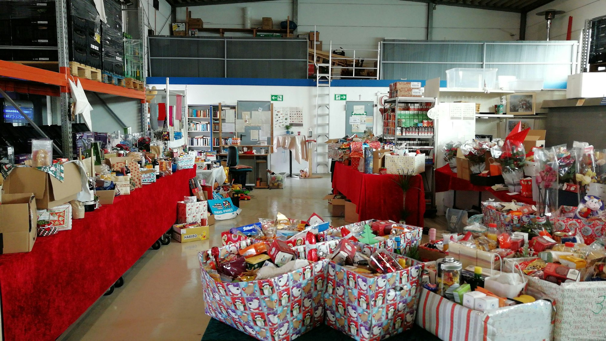 In der Halle der Zülpicher Tafel stehen viele weihnachtlich-dekorierte Kiste mit allerhand Plätzchen, Backzutaten und anderen Leckereien gefüllt.