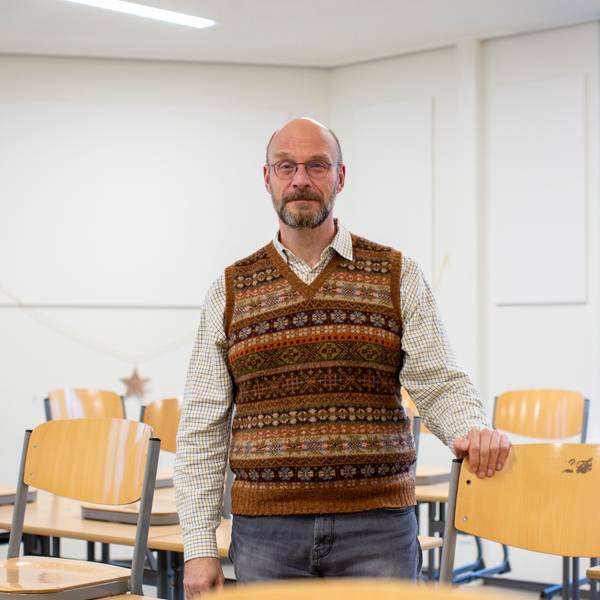 Ein Lehrer mit Strickpullunder steht in einem Klassenzimmer.