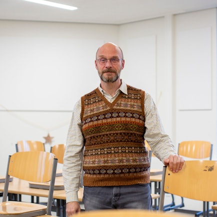Ein Lehrer mit Strickpullunder steht in einem Klassenzimmer.