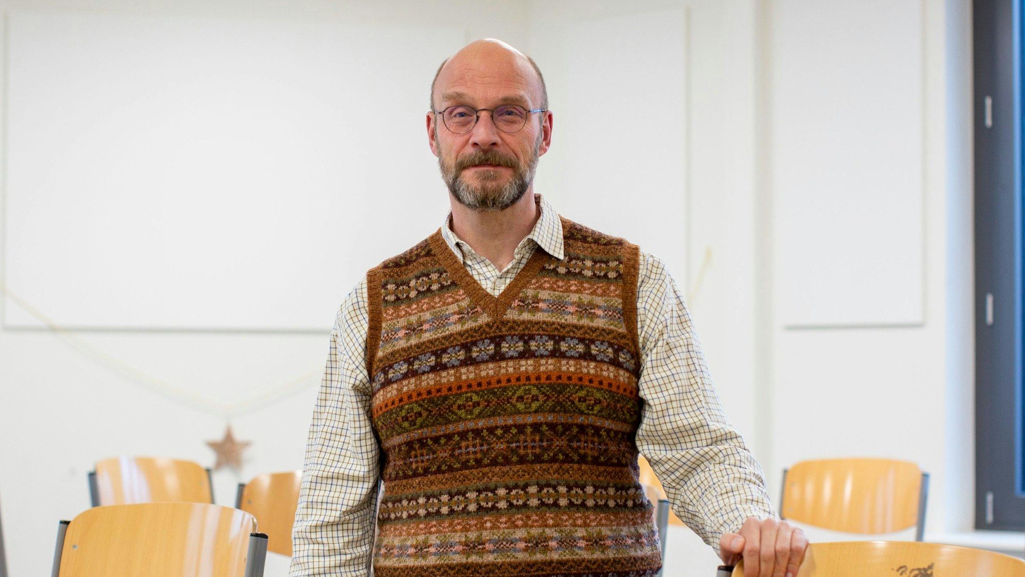 Ein Lehrer mit Strickpullunder steht in einem Klassenzimmer.