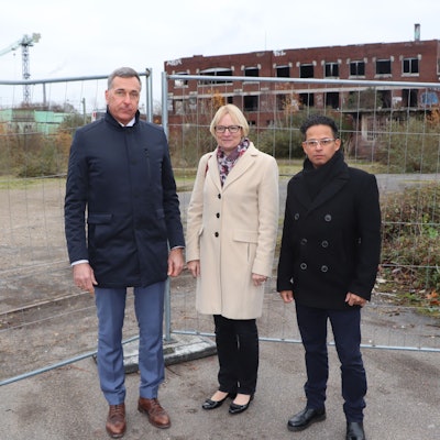 Auf dem Foto sind Wesselings Bürgermeister Ralph Manzke, KVB-Vorstandsvorsitzende Stefanie Haaks und Projektleiter Manuel Franke (v.l.) vor dem Bauzaun der Industriebrache der ehemaligen Norton-Fabrik in Wesseling am Kronenweg zu sehen.