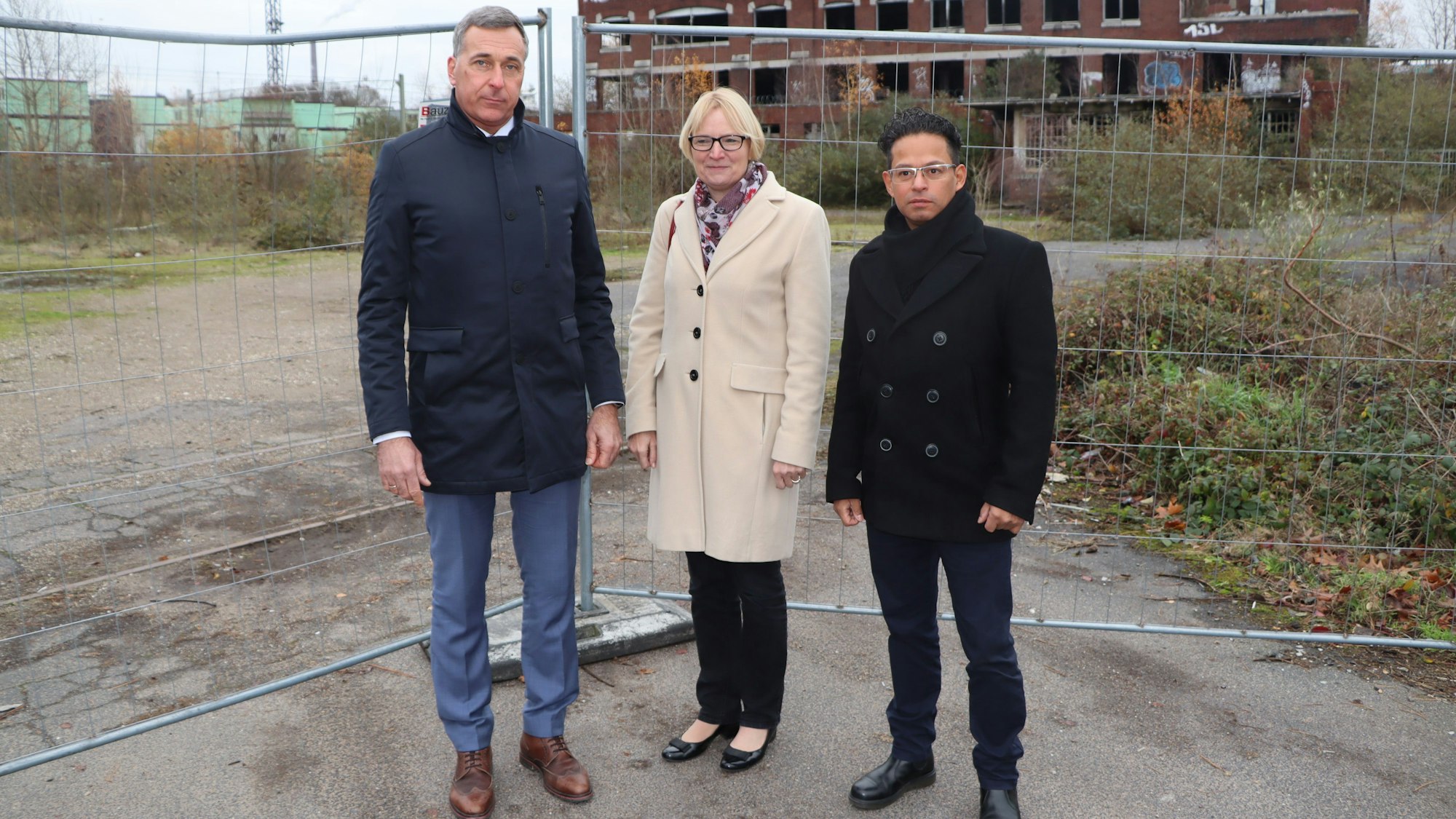 Auf dem Foto sind Wesselings Bürgermeister Ralph Manzke, KVB-Vorstandsvorsitzende Stefanie Haaks und Projektleiter Manuel Franke (v.l.) vor dem Bauzaun der Industriebrache der ehemaligen Norton-Fabrik in Wesseling am Kronenweg zu sehen.