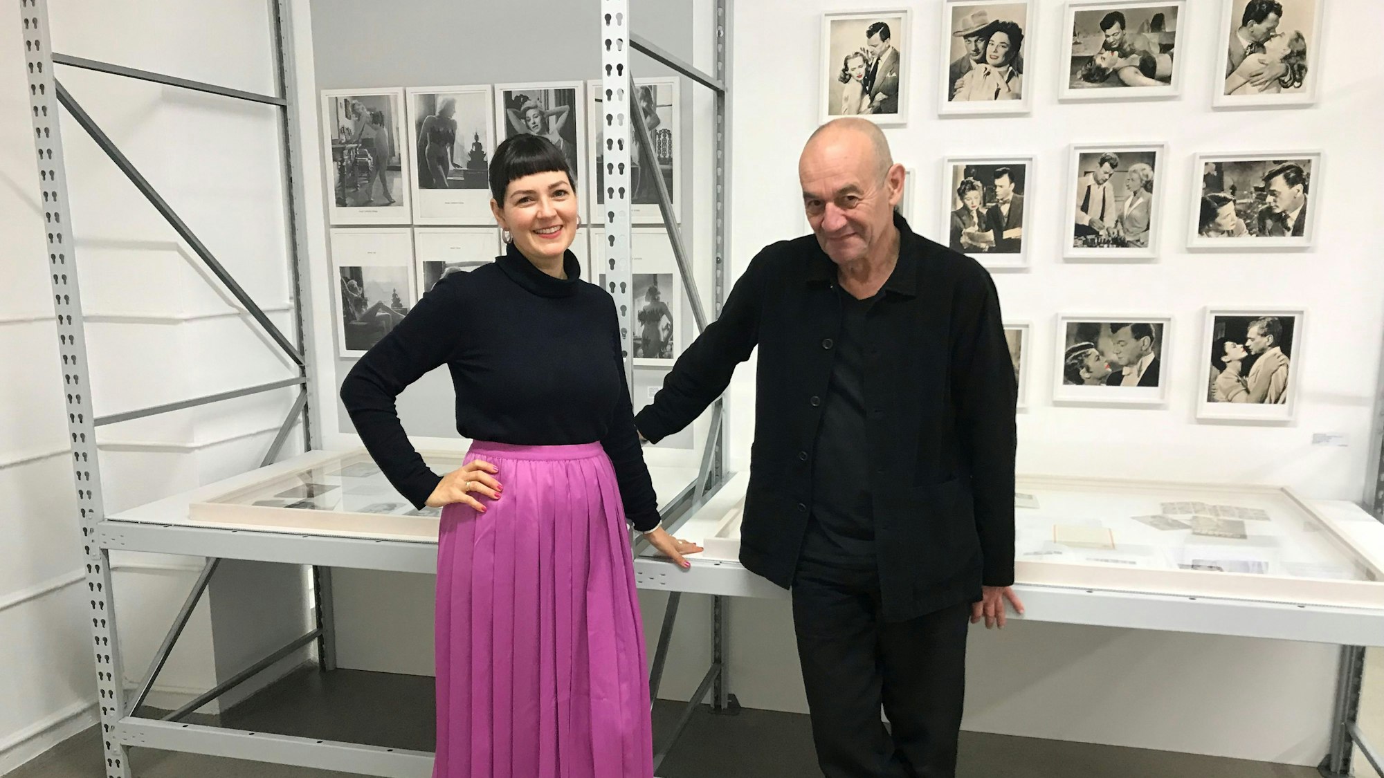 Eine Frau und ein Mann stehen vor einem Regal. Hinter ihnen hängen viele Fotos in Schwarz-Weiß.