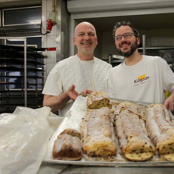 Zwei Bäcker halten ein riesiges Backblech in ihren Händen, darauf liegen vier Christstollen.